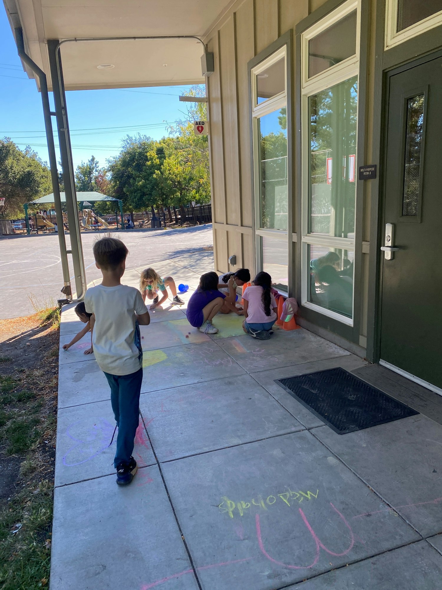 Children playing with sidewalk chalk outside a school building under a sunny sky.