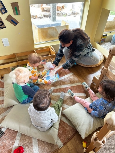 A woman reads to four toddlers seated on cushions in a cozy room with a window and wooden furniture.