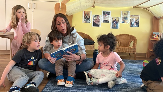 A woman reading a book to young children in a cozy classroom setting, with kids gathered around her.