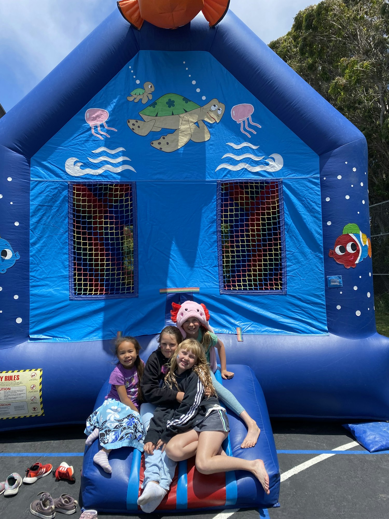 A colorful inflatable bounce house with ocean-themed graphics, featuring four children sitting on it, enjoying their time outdoors.