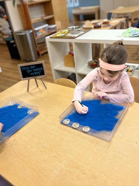 Child drawing in blue sand tray, small sign asking "How are you feeling today?" nearby in a classroom setting.
