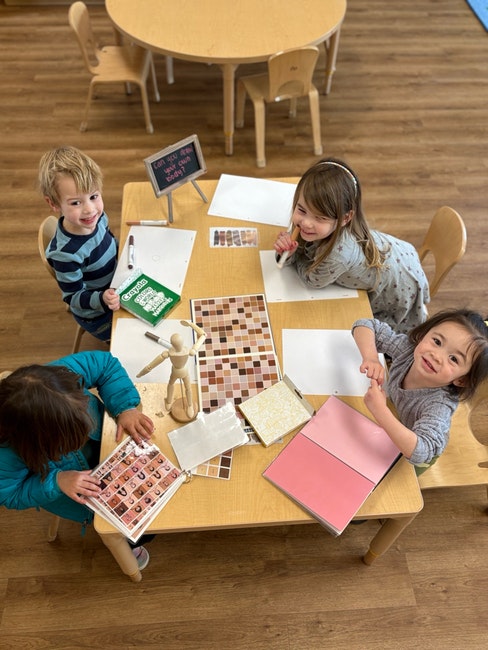 Four children sitting at a table with paper and art materials, smiling and drawing.