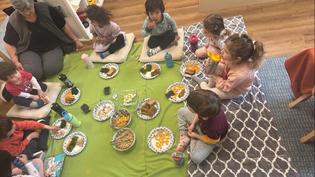 Children and an adult seated around a picnic with various dishes on a green blanket indoors.
