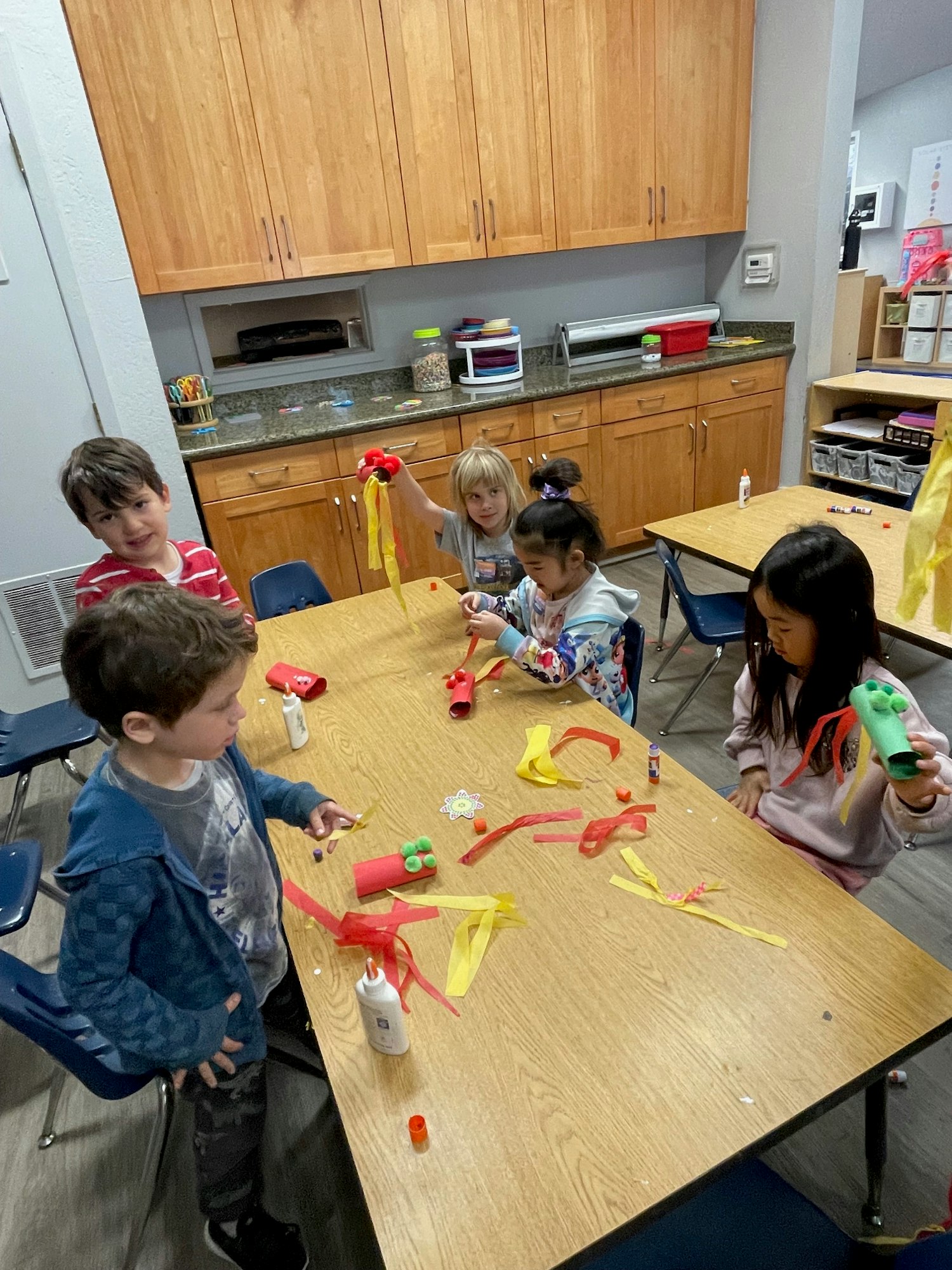Children crafting with colored paper and glue around a table in a classroom.