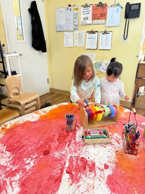 Two children play with art supplies on a table covered in red and orange paint in a classroom setting.