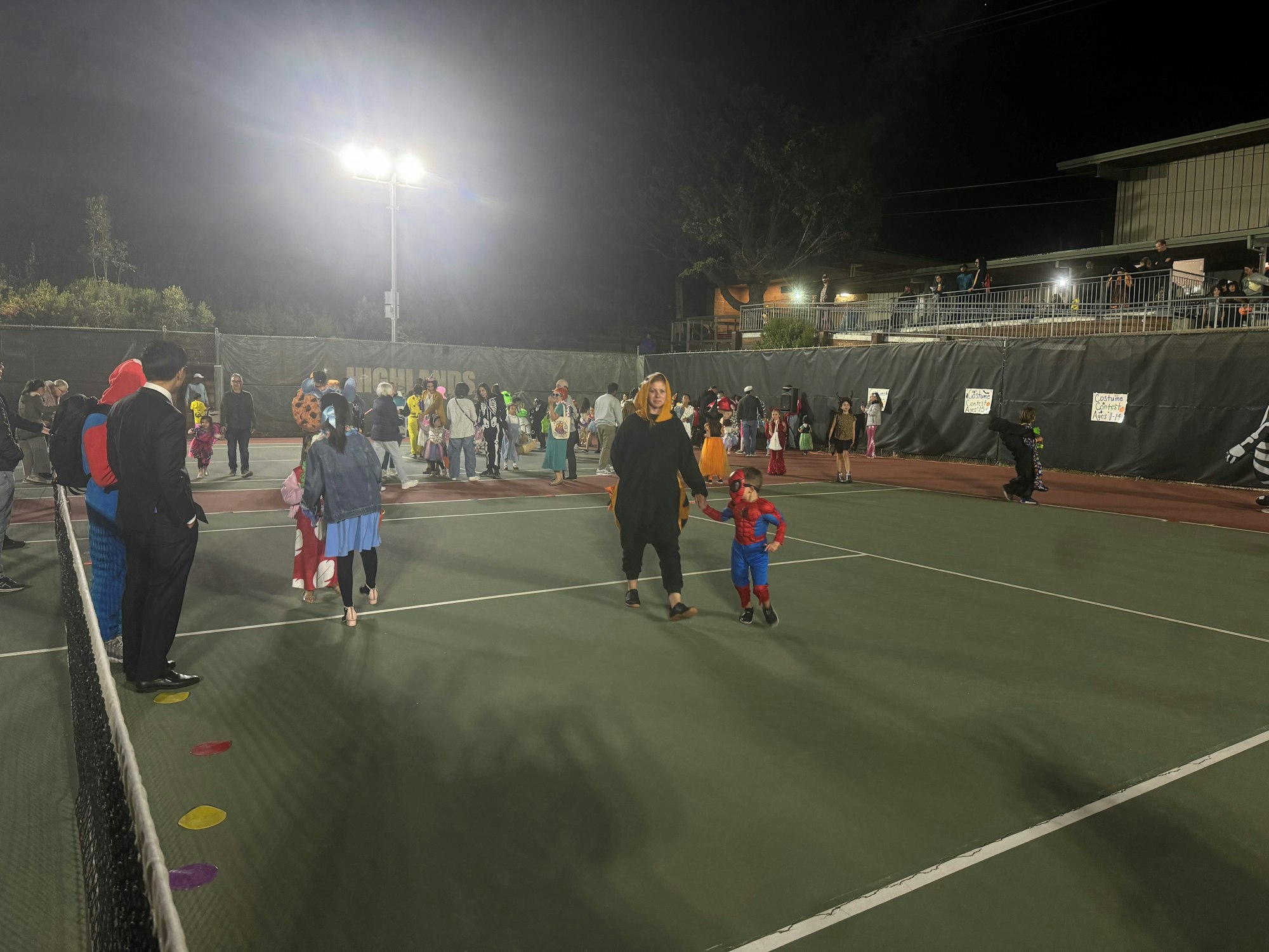 People in costumes at an outdoor night event, likely a Halloween party on a tennis court.
