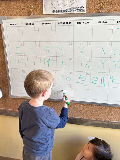 A child writes on a calendar whiteboard with a green marker, while another child watches.