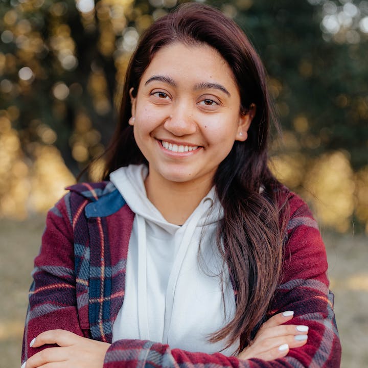 A person smiling outdoors, wearing a red plaid jacket over a white hoodie, with trees blurred in the background.