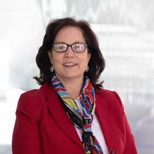 A woman in a red blazer and colorful scarf, wearing glasses, standing indoors.