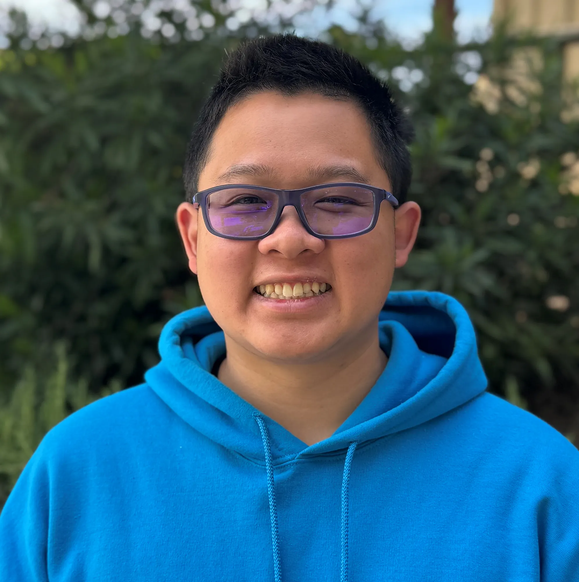 A smiling person wearing a bright blue hoodie stands outdoors with greenery and a clear sky in the background.