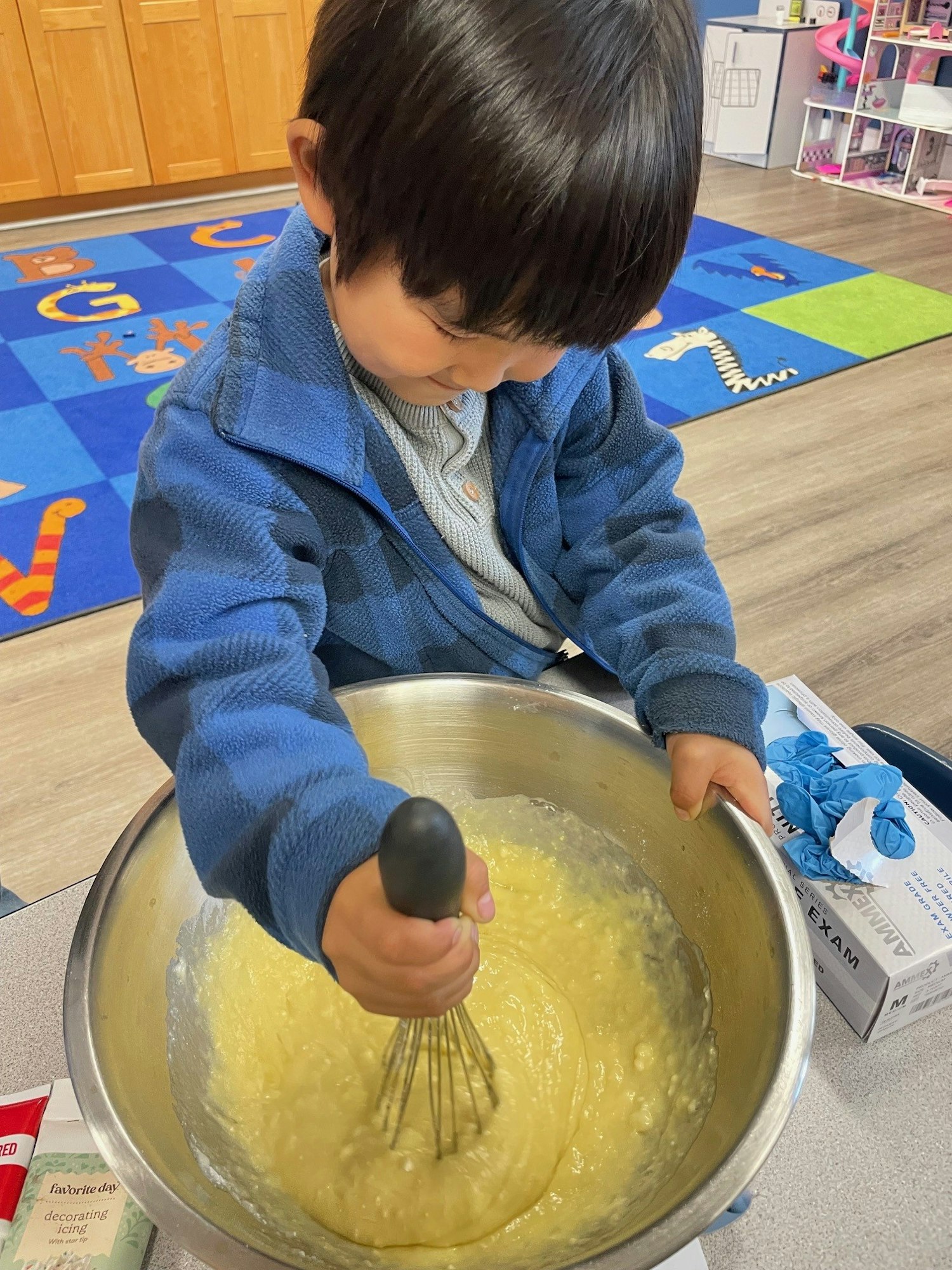 A young child is stirring batter in a mixing bowl with a whisk, focused on their task in a colorful play area.
