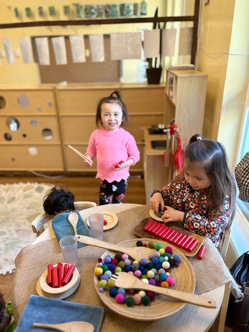 Children playing with colorful balls and small musical instruments at a table in a classroom setting.