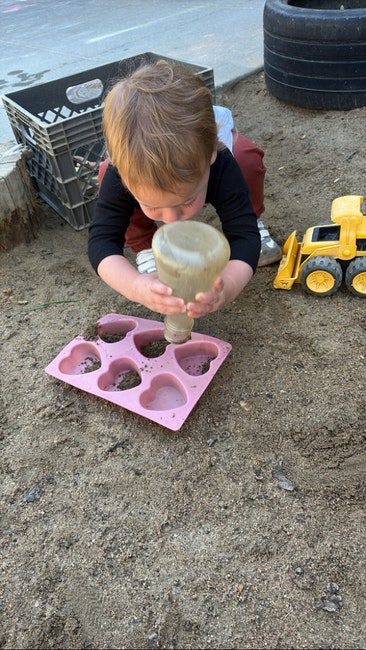 Child playing in sand with bottle, heart-shaped mold, and toy truck.