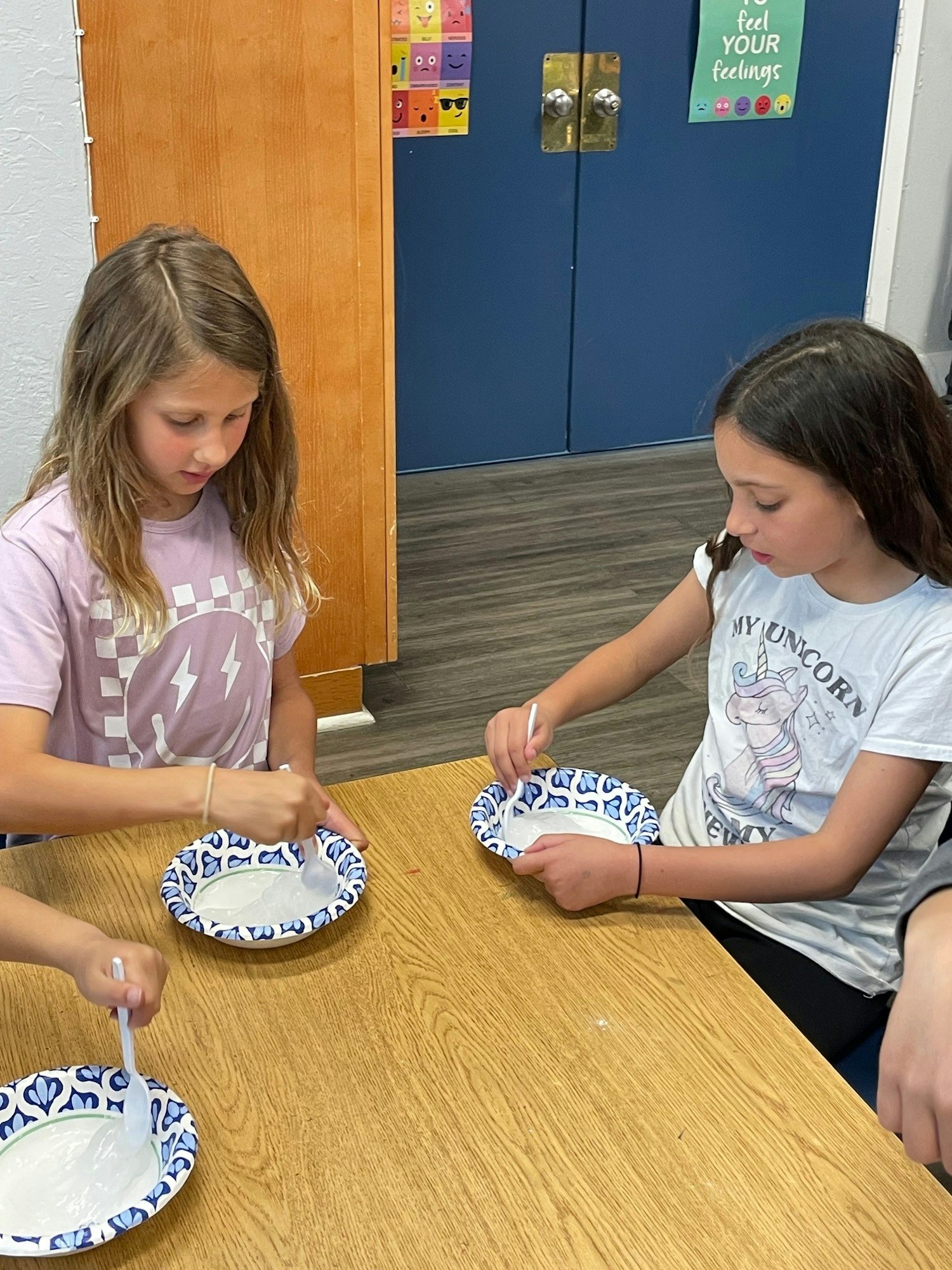 Two children sitting at a table, stirring white substance in paper plates with plastic spoons.