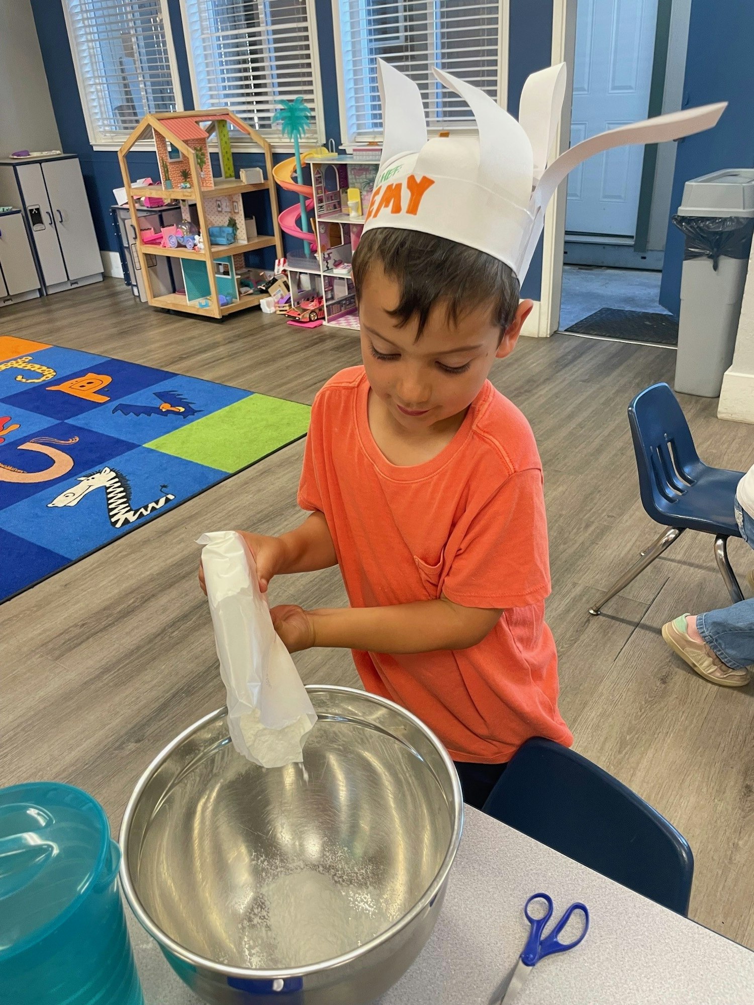 A child in an orange shirt wearing a paper hat is pouring flour into a metal bowl in a playroom.