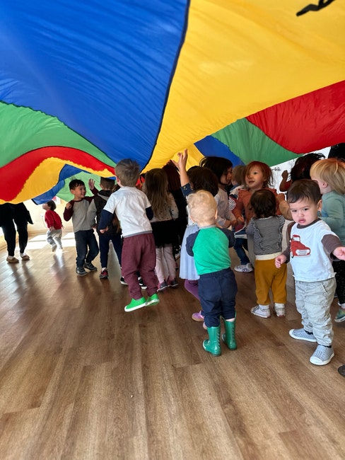 Children playing under a colorful parachute indoors on a wooden floor.