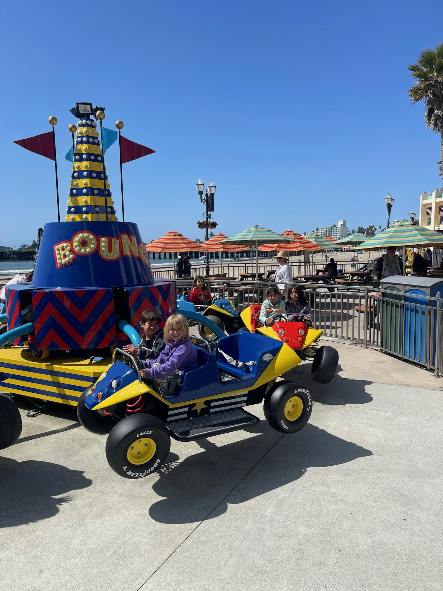 Children on a colorful amusement park ride with a blue tower and umbrellas in the background.