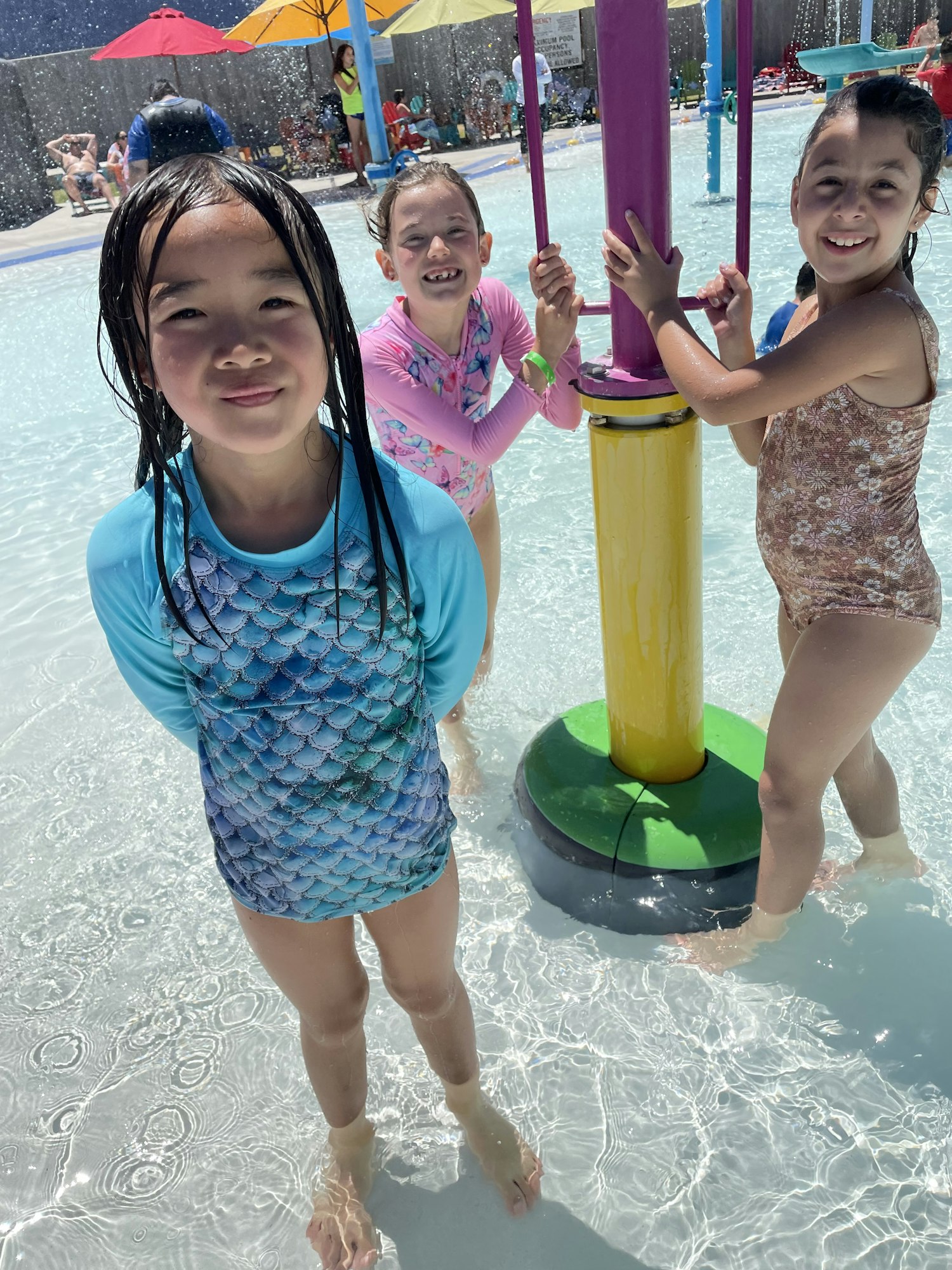 Three girls are enjoying a splash pad on a sunny day, smiling and playing in shallow water surrounded by colorful umbrellas.