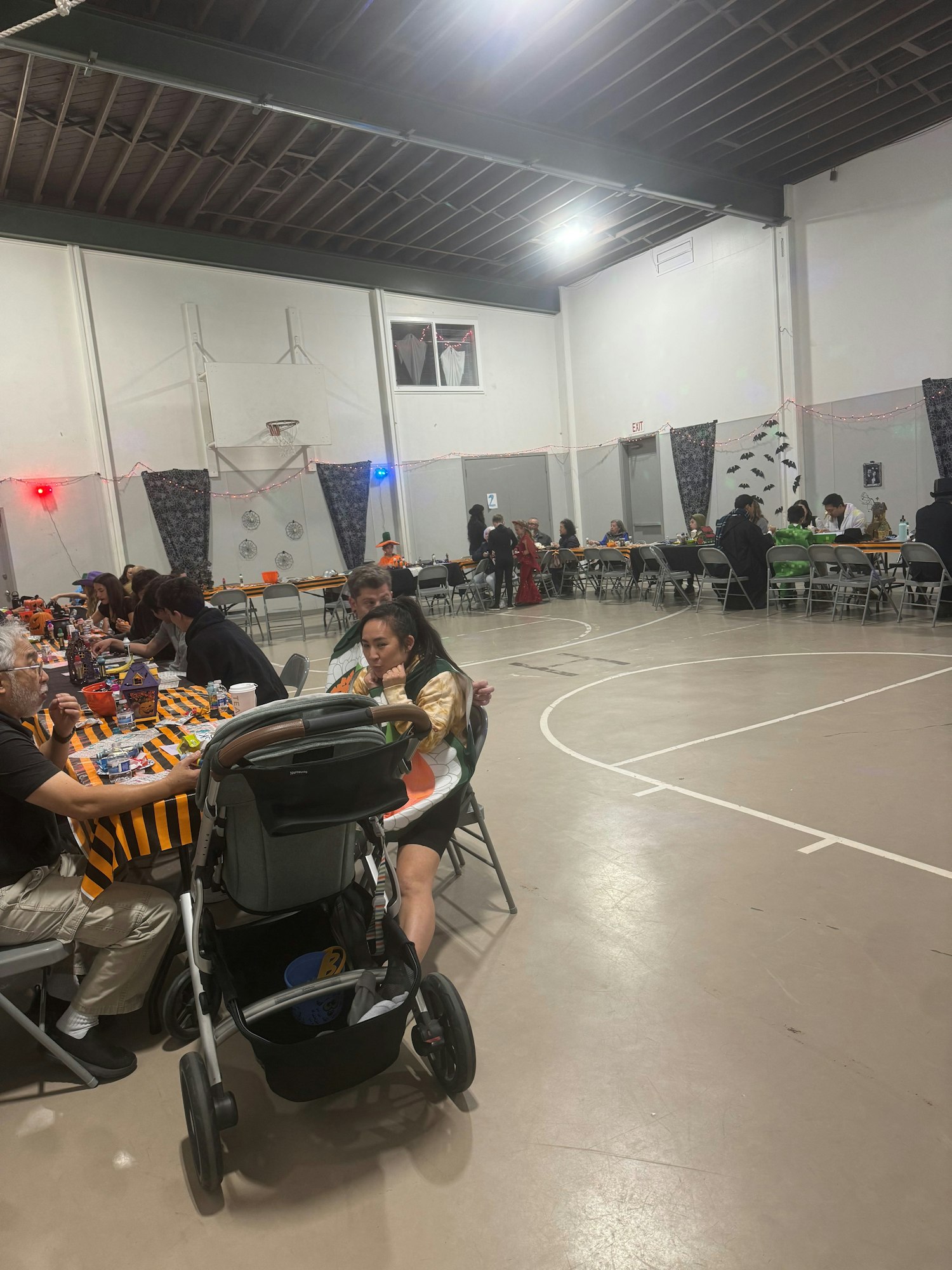 Indoor gathering with people sitting at tables, a stroller in the foreground, and sports court lines on the floor.