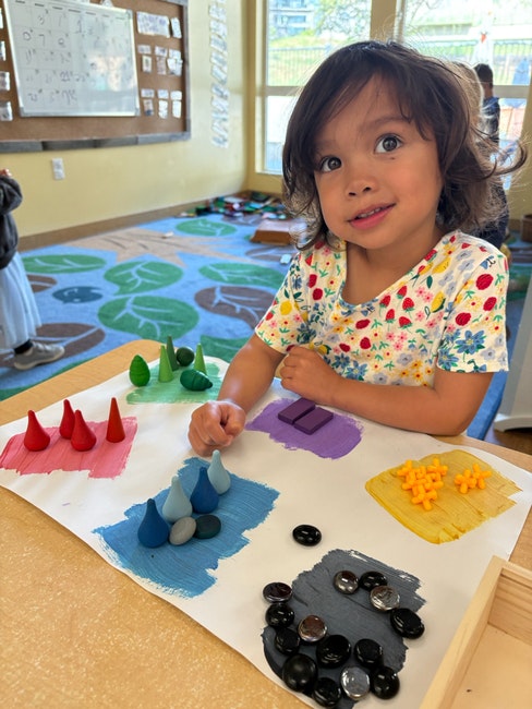 A child in a classroom, smiling, with colored cones and stones on a paper with painted sections.