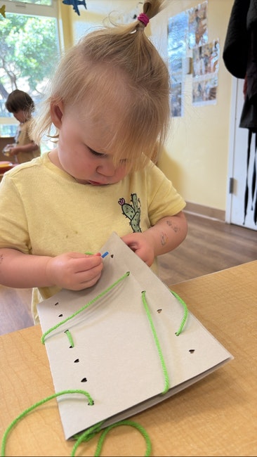 A child is focused on a lacing activity using green yarn and a perforated cardboard.
