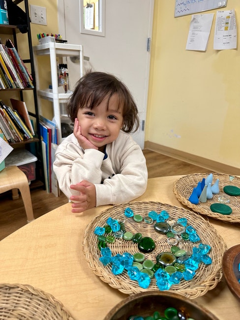 A child smiles at a table with blue and green stones on woven trays in a classroom.
