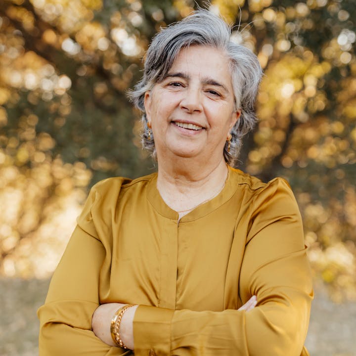 Older woman smiling, wearing a mustard blouse, arms crossed, standing outdoors.