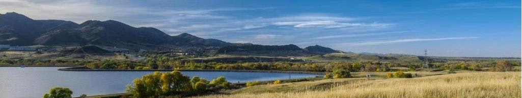 A scenic view of a lake surrounded by grasslands and distant mountains under a blue sky.
