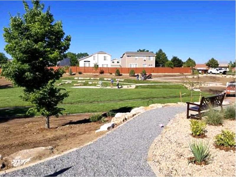 A landscaped garden with a pathway, bench, plants, and people in the distance against a backdrop of houses and clear blue sky.