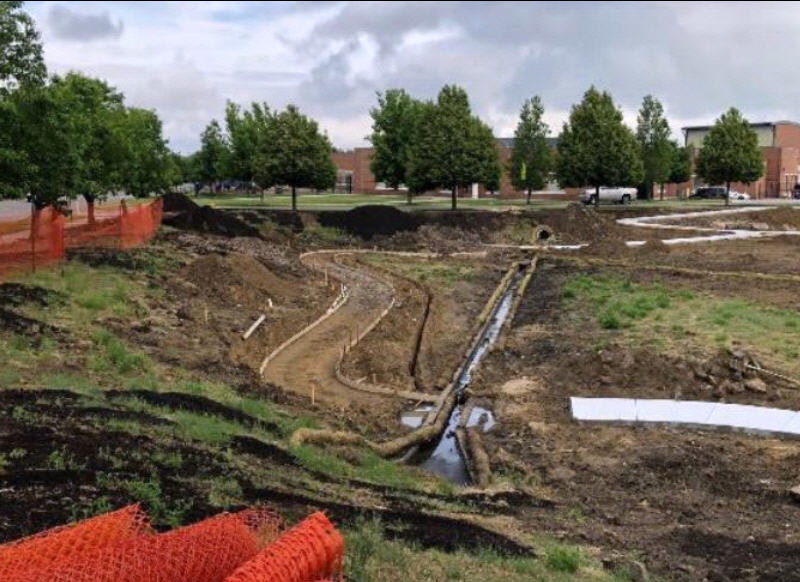 Construction site with dug up earth, pipes exposed, buildings in the background, and orange safety fence.