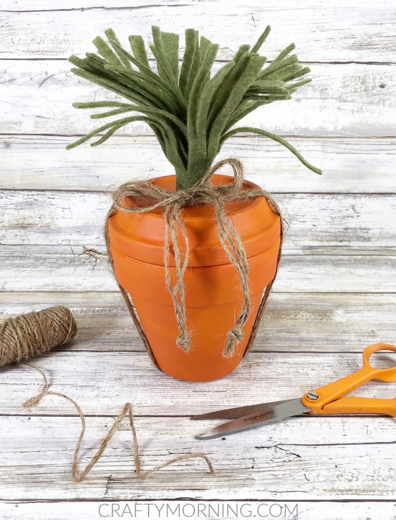 A carrot-shaped craft with green felt leaves, twine tied around, scissors, and a roll of twine on a wooden surface.