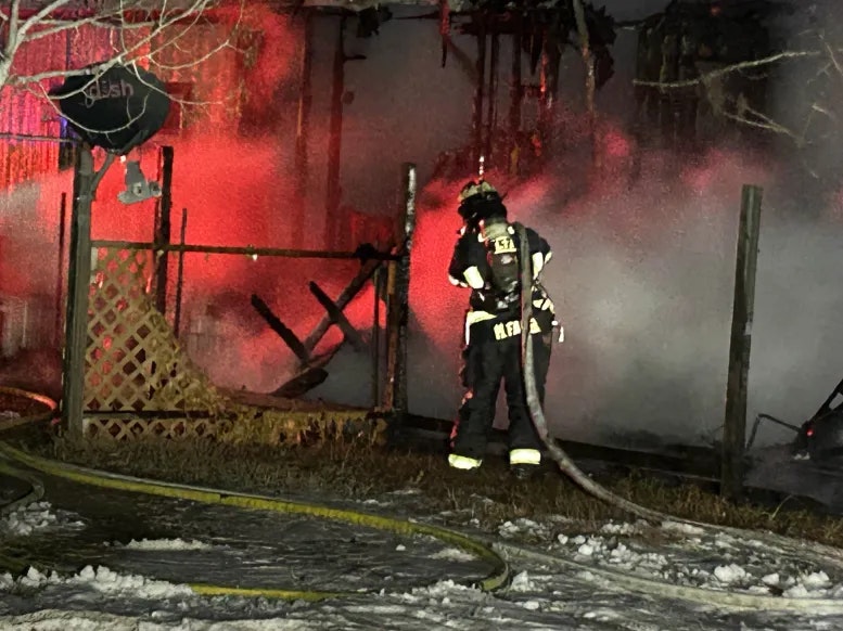 A firefighter near a burnt structure with hoses, red lights, and snow on the ground.