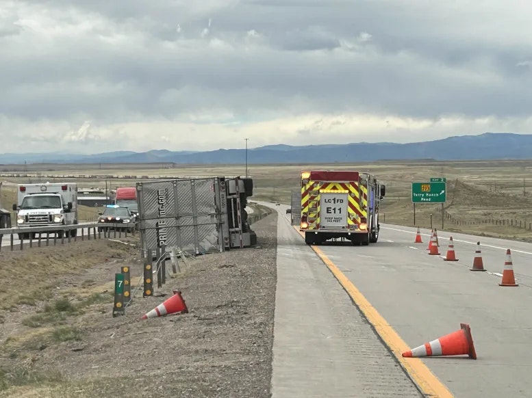 Overturned semi-truck on highway, emergency services on site, with traffic cones and grassland in the background.