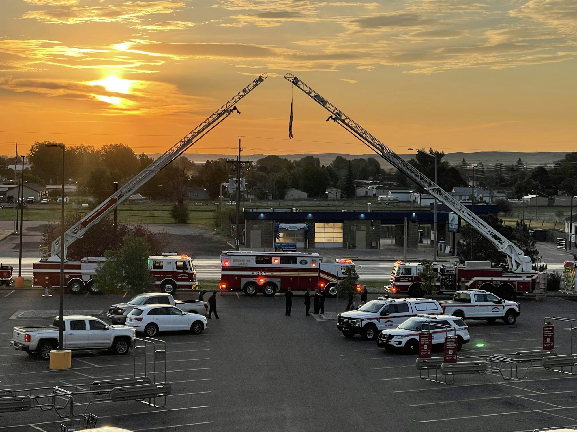 Fire trucks extending ladders across a sunset sky, police cars, and onlookers in a parking lot.