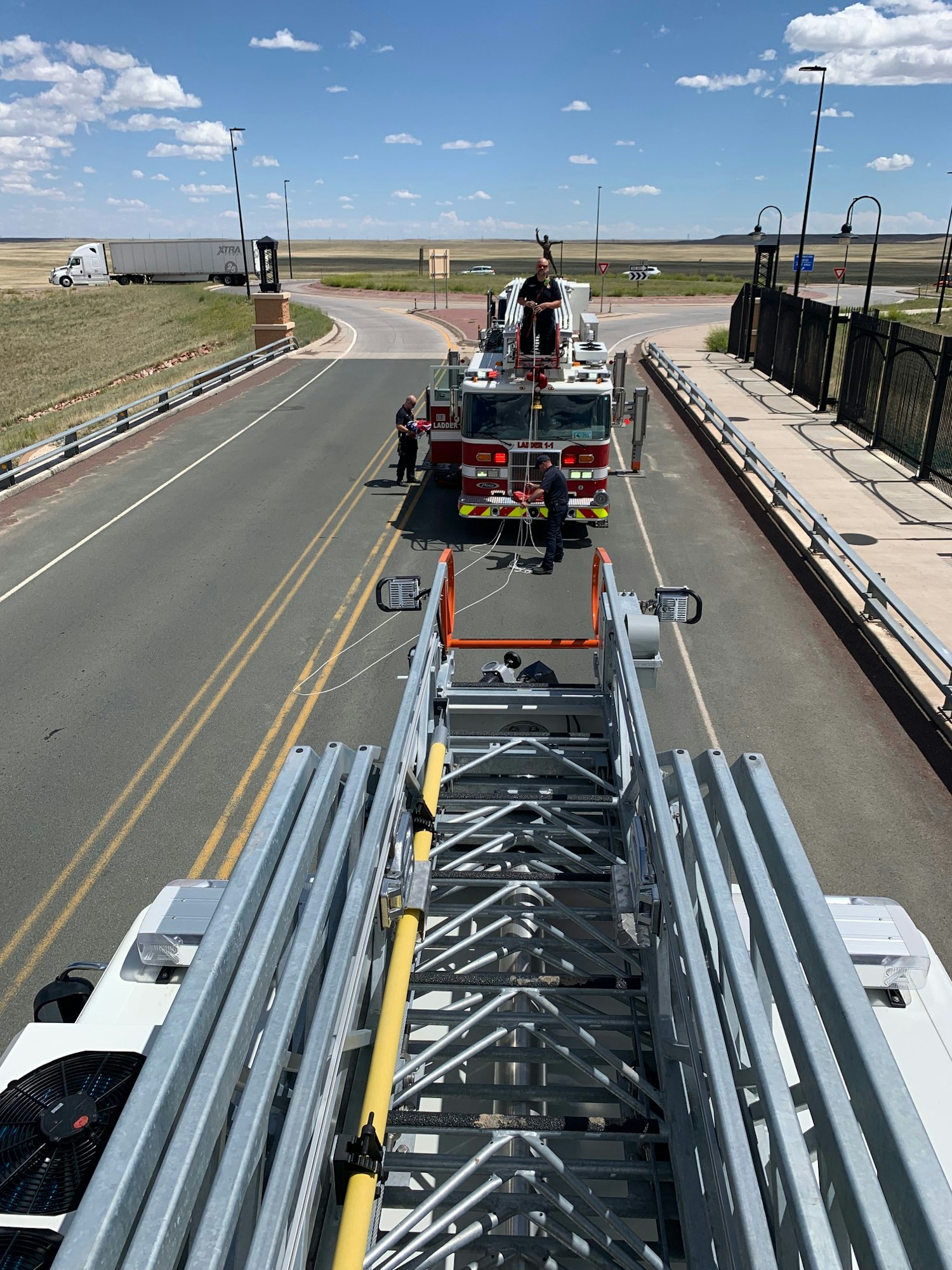 Fire truck on the road with extended ladder, people working on it, clear sky and some vehicles seen in the background.