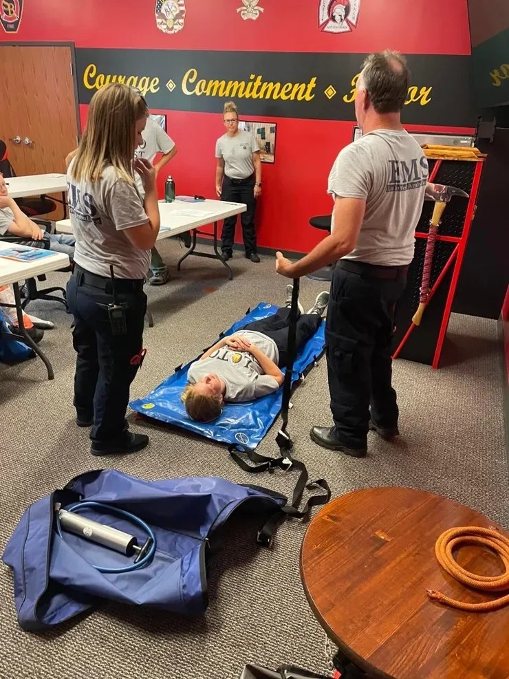 People in an EMS training room; one person lies on a blue stretcher.
