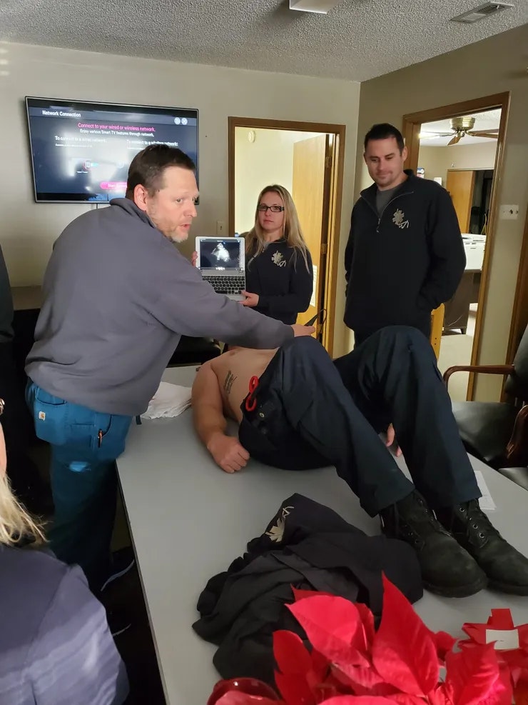 A person lies on a table as others watch a laptop screen, possibly during a medical demonstration or training session.