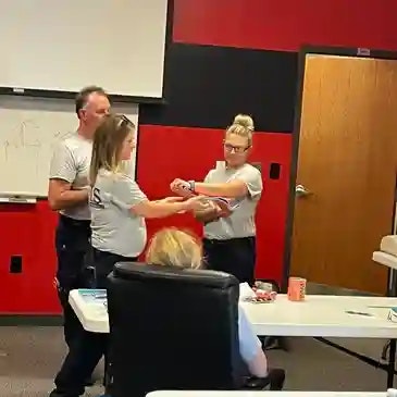 Three people are demonstrating a medical technique in a classroom setting, observed by a seated person.