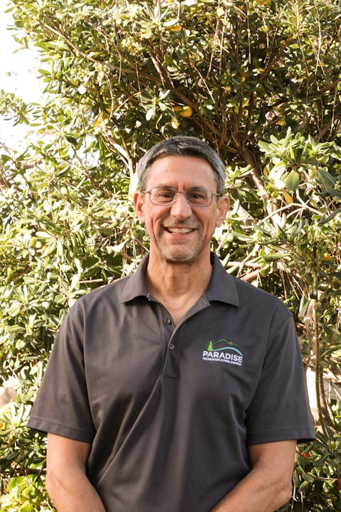 A smiling man in a branded polo shirt standing in front of green foliage.