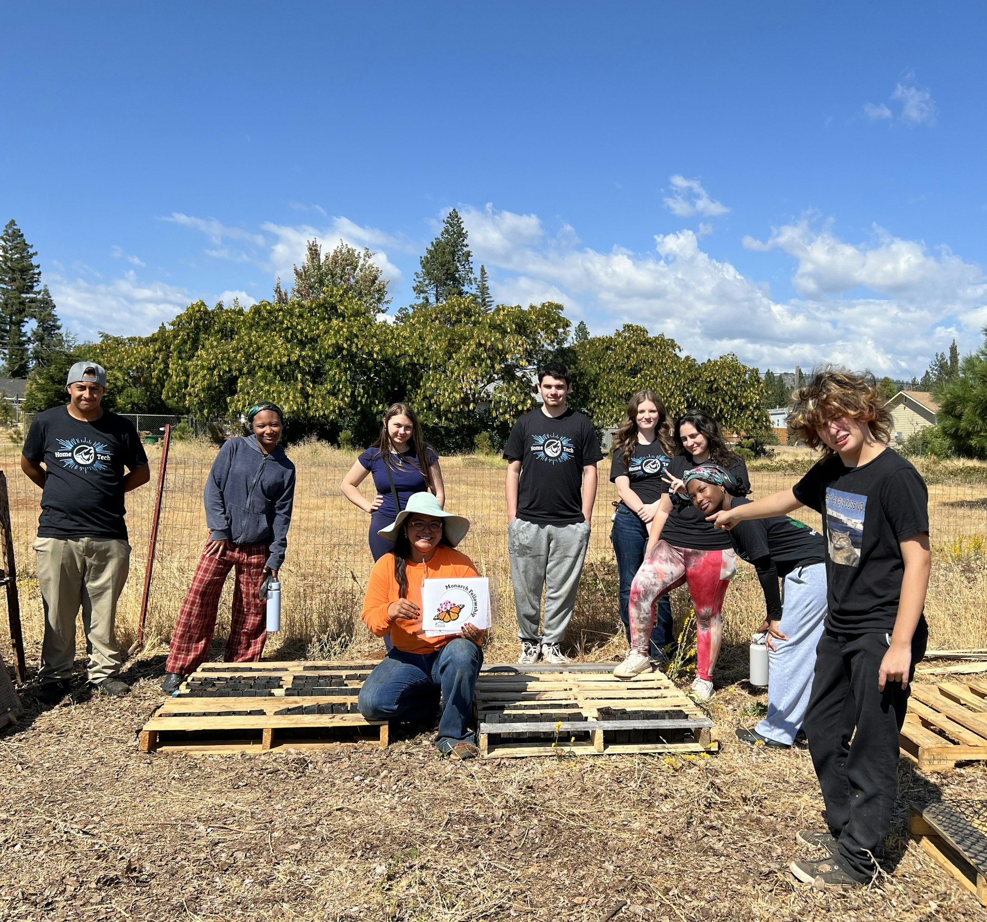 A group of people poses outdoors, some holding a sign, in a field with trees and sunny skies in the background.