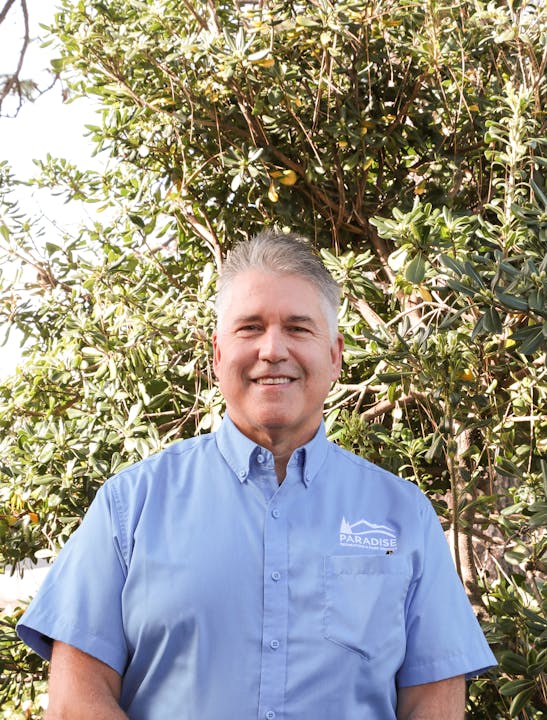 A smiling person in a blue shirt with "PARADISE" logo, standing in front of a leafy background.