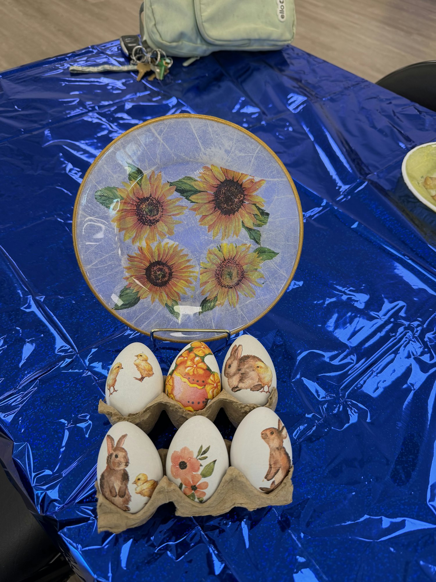 The image features a blue tablecloth, a decorative plate with sunflowers, and six decorated eggs in a carton.