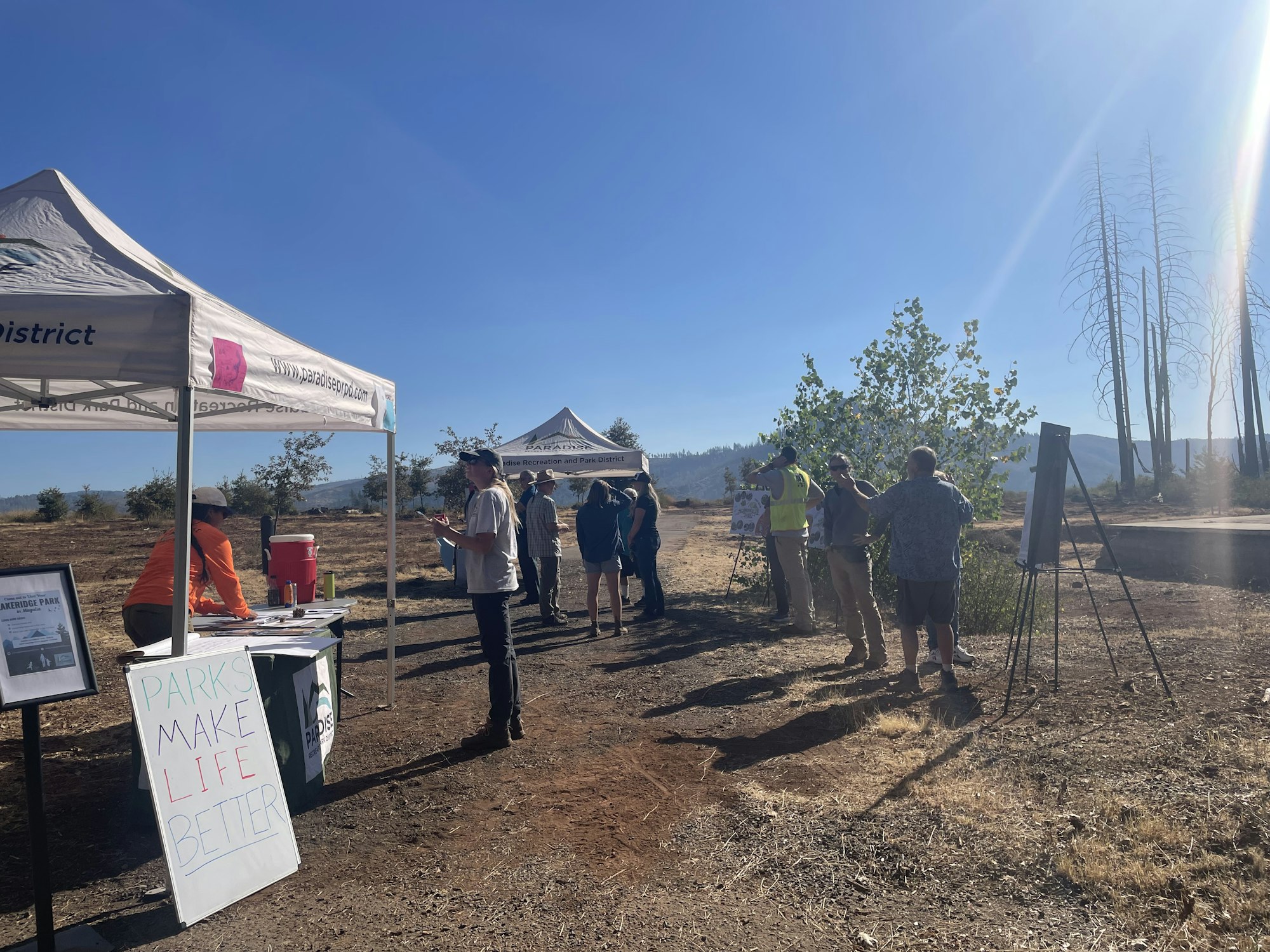 A group of people gather under small white canopy tents at an outdoor community event on a clear, sunny day. Informational displays and a sign-in table are set up along a dirt path, with trees and a mountain range visible in the distance.