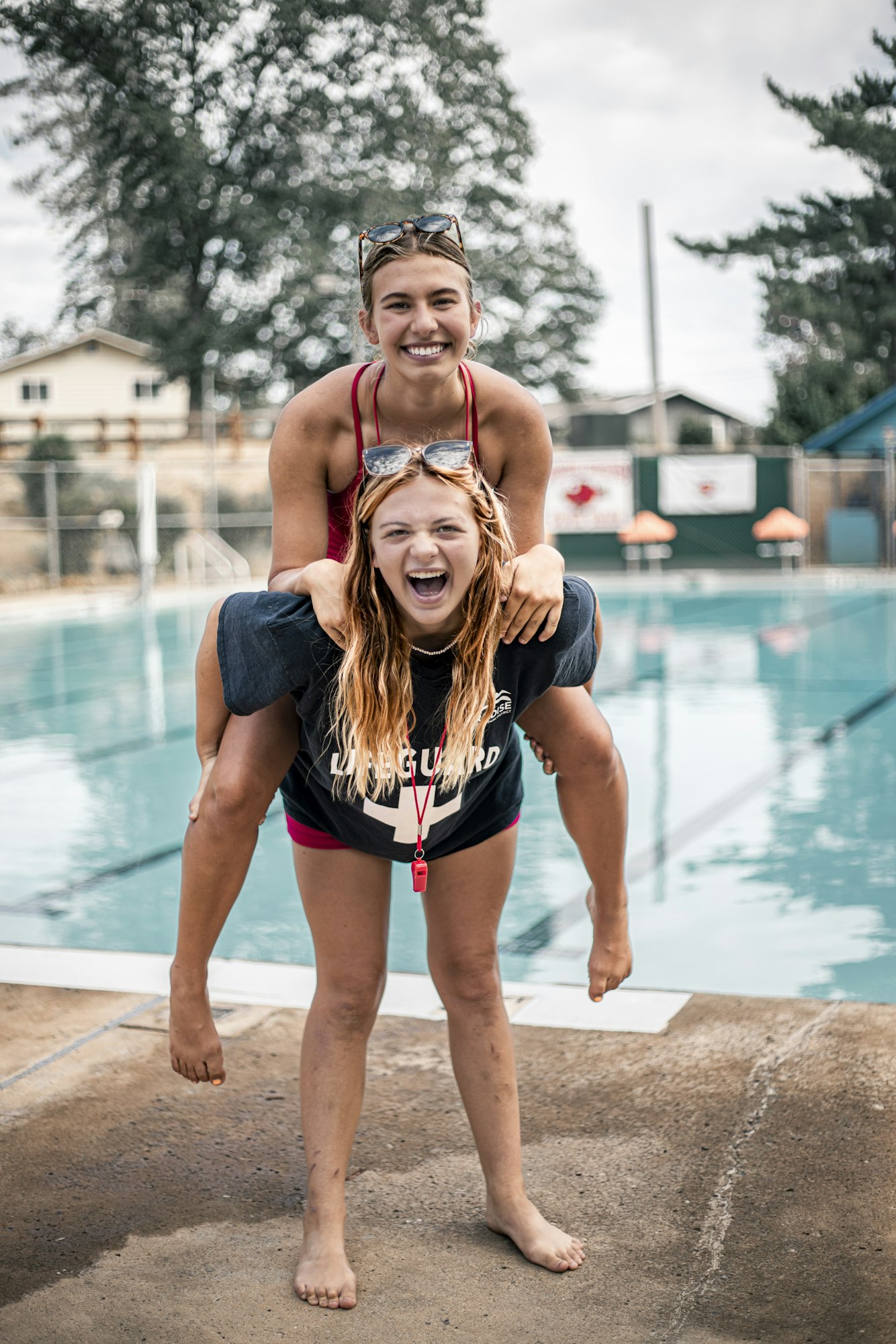 Two lifeguards having fun, one giving the other a piggyback ride by a poolside, both smiling and wearing sunglasses.