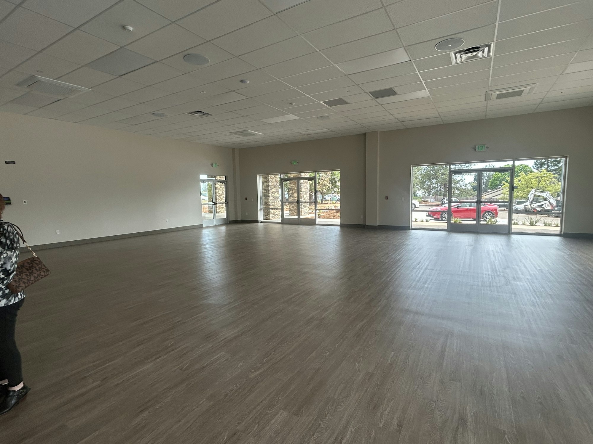 An empty room with wooden floors and large windows, doors on the side, and a person partially visible.