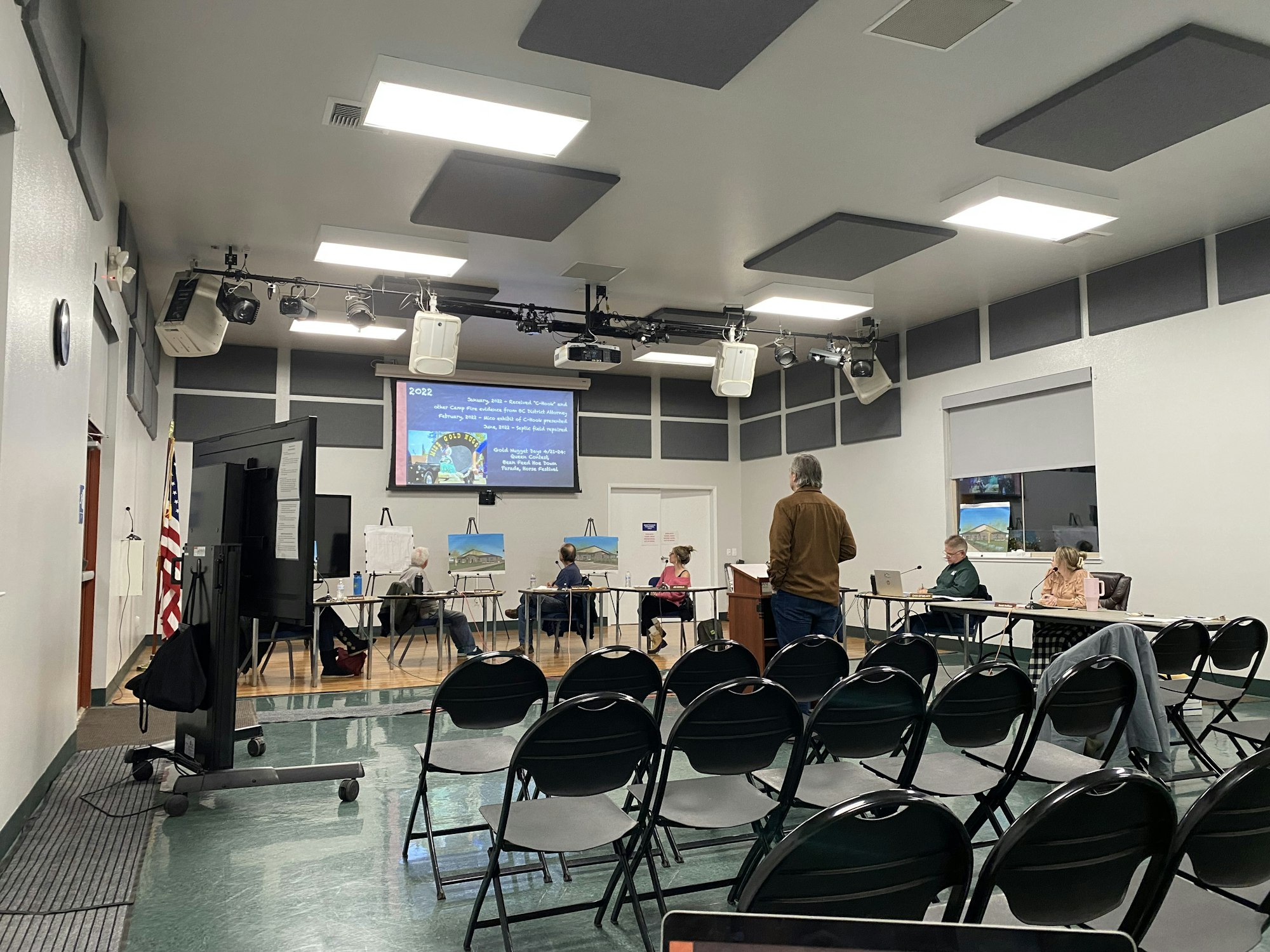 An indoor meeting room with people at tables, a presenter, empty chairs, a screen displaying a presentation, and an American flag.
