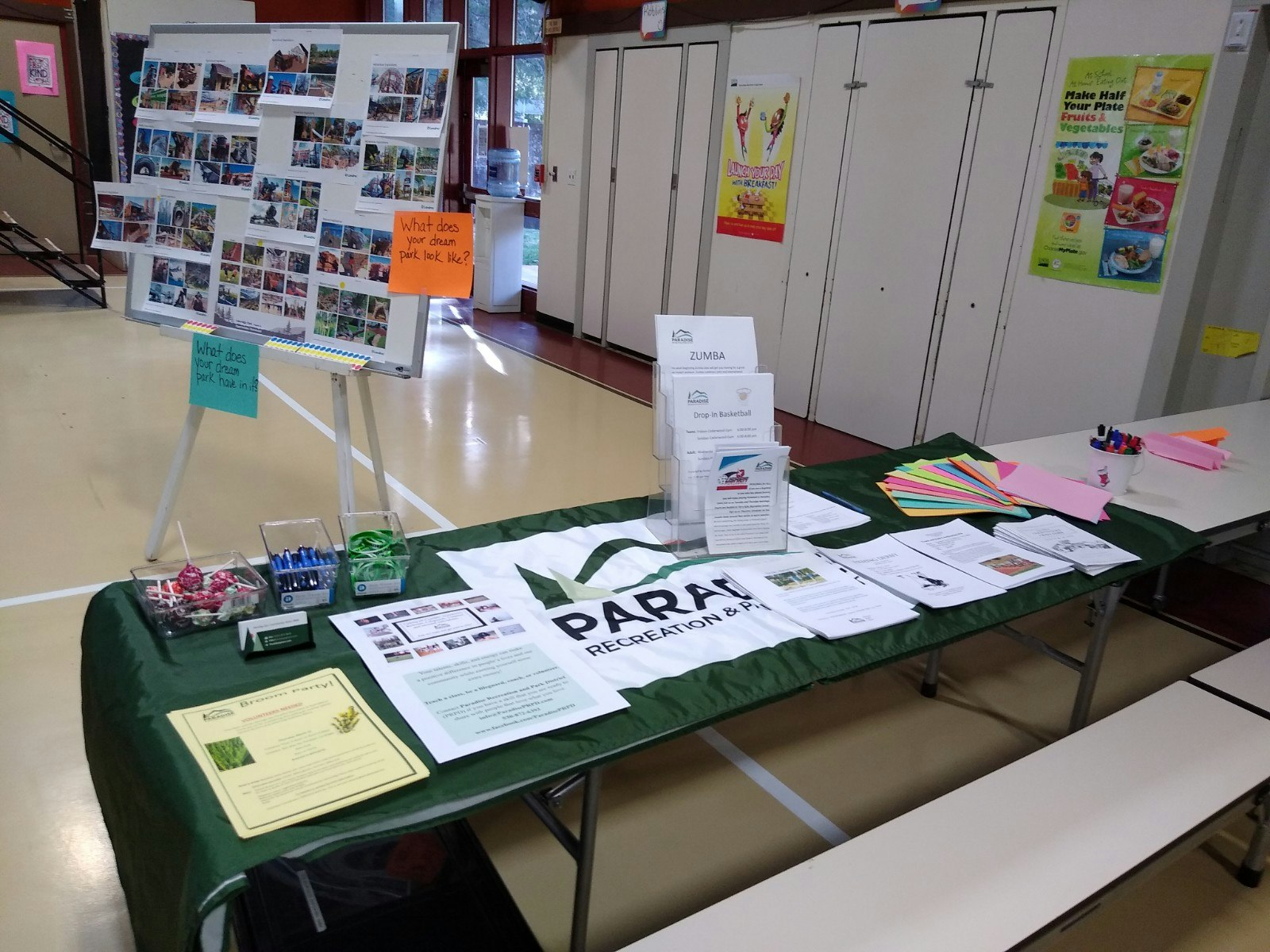 The image shows a community event table with informational brochures, a display board asking about dream parks, and activity promotions.