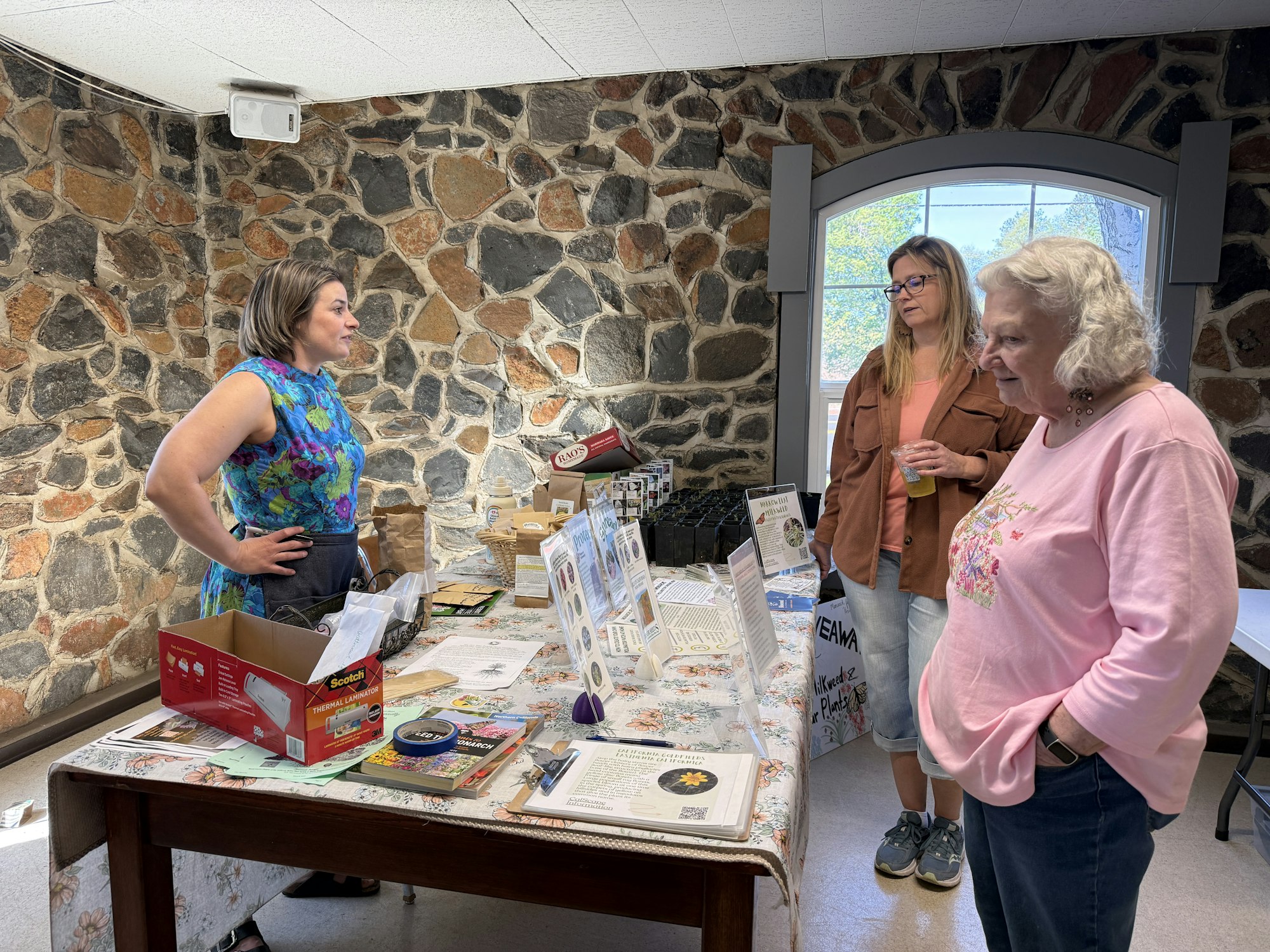 Three women are engaging at a table with various materials in a stone-walled room, possibly discussing a local event or products.