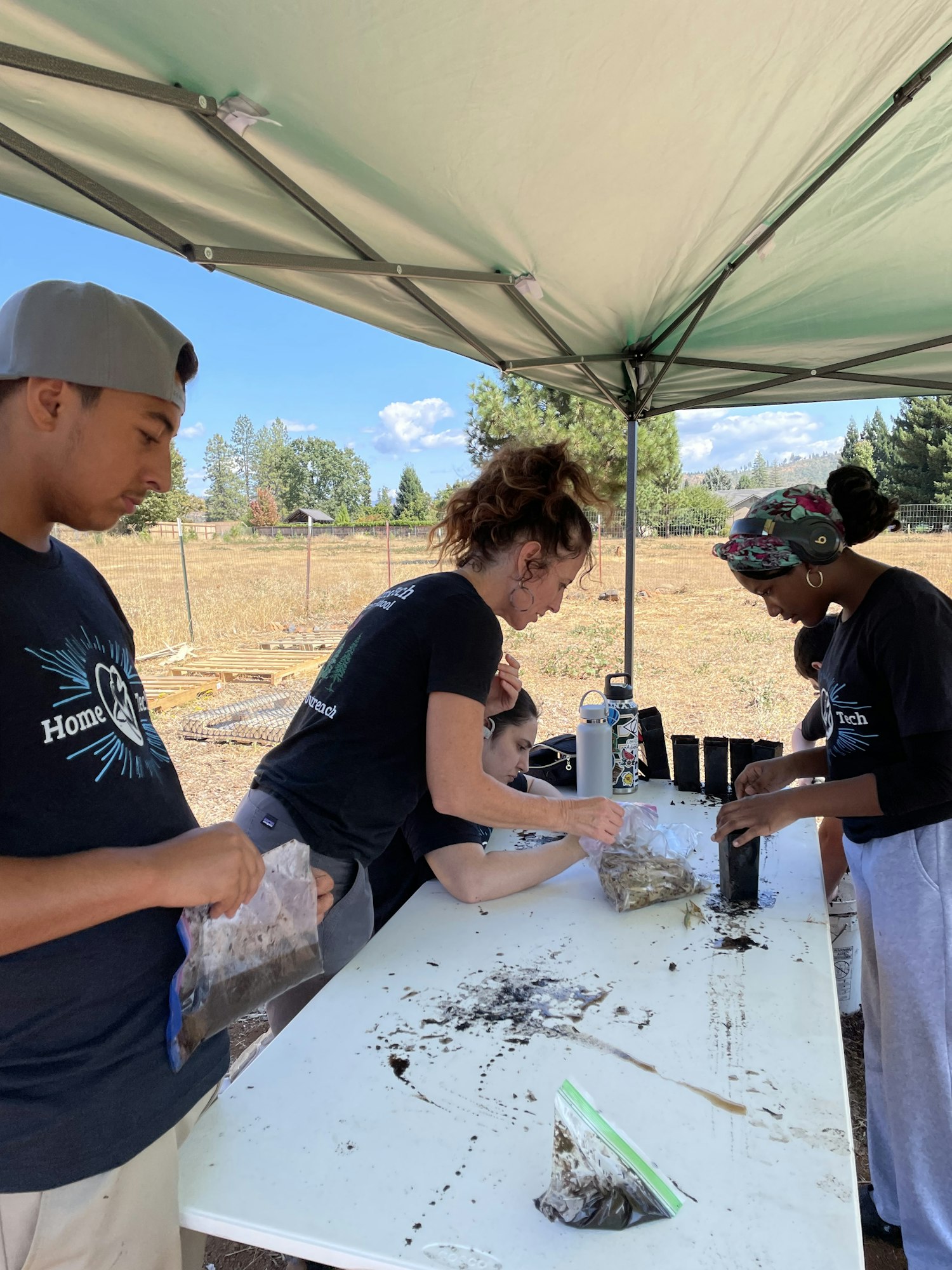 People working outdoors at a table, handling soil and planting materials under a tent on a sunny day.