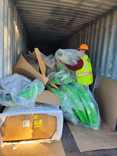 A person in safety gear is sorting through green plastic items and cardboard boxes inside a shipping container.